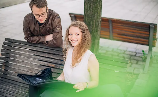 Sophie Buchta sitzt auf einer Bank im Freien und lächelt in die Kamera, während Jan-Michael Bussmann an die Bank gelehnt ist und konzentriert auf eine aufgeschlagene Mappe blickt.