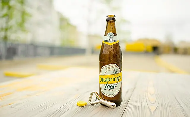 Geöffnete Flasche Ottakringer Lager Bier mit Kronkorken und Flaschenöffner auf einem Holztisch im Freien.