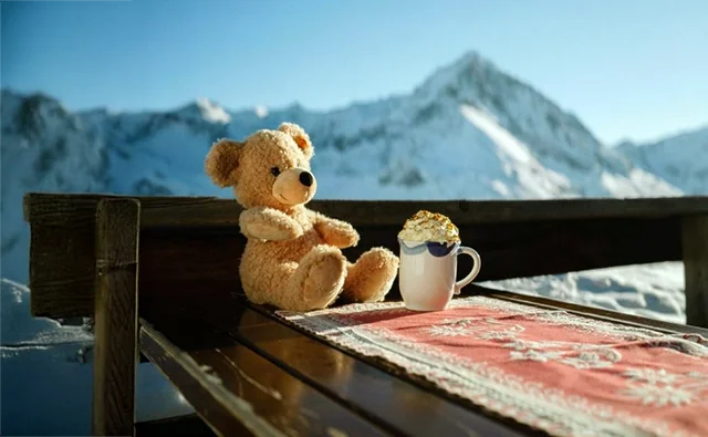 Szene aus der zielgruppenspezifischen Werbekampagne 'Immer für dich da'. Steiff-Teddybär Pauli mit heisser Schokolade auf einer Almhütte vor imposanter Alpenkulisse.
