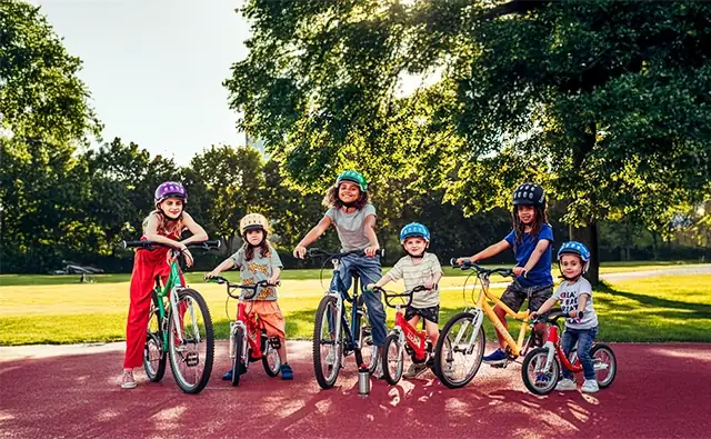 Sechs Kinder verschiedenen Alters und Hautfarbe auf farbenfrohen Woom-Fahrrädern in einem sonnigen Park. Symbolbild für die starke Markenpräsenz und globales Wachstum in verschiedenen Märkten.