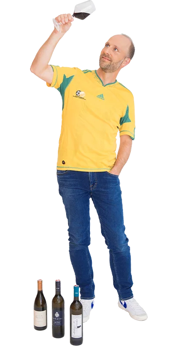 Dr. Christian Bosch steht mit legerer Kleidung und vier Weinflaschen vor sich, während er ein Glas Rotwein prüfend gegen das Licht hält - Geniesser mit Fachwissen.