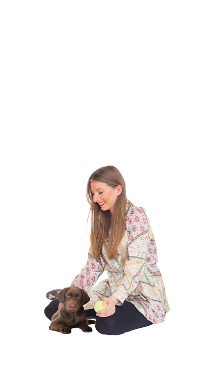 Caroline Peinitz-Kichler, MSc kniet mit einem kleinen Labradorwelpen am Boden, eine gelbe Tennisball in der Hand - tierlieb und herzlich.