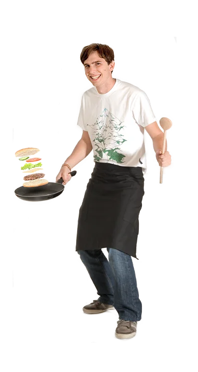 Manuel Nussbaumer, MSc (WU) trägt eine Kochschürze, jongliert einen gestapelten Burger in einer Pfanne und hält in der anderen Hand einen Kochlöffel - kreativ, verspielt und kochbegeistert.