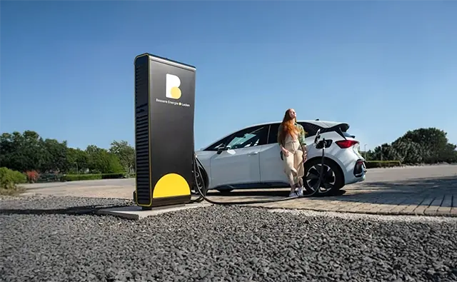 Eine Kundin lehnt an einem Elektroauto, das an einer modernen Ladesäule der Burgenland Energie angeschlossen ist, und sonnt sich entspannt, während sie auf den Ladevorgang wartet.