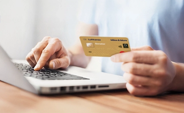 Eine Person hält eine goldene Lufthansa Miles & More-Kreditkarte in der Hand und tippt mit der anderen Hand auf einem Laptop.