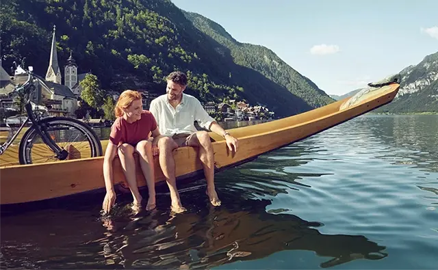 Paar entspannt auf einem traditionellen Holzboot im Hallstätter See mit Blick auf die Alpen. Darstellung der idyllischen österreichischen Sommerlandschaft für touristisches Marketing.