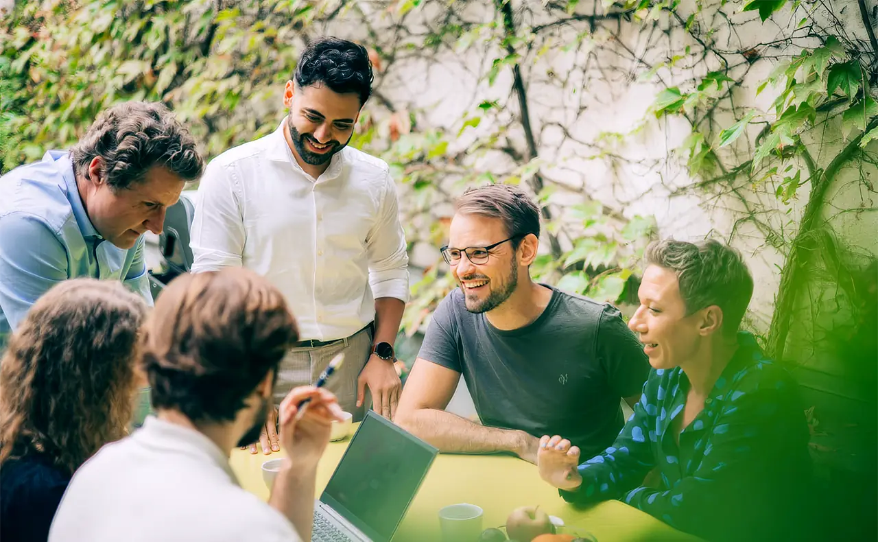 Gereon Friederes, Philipp Zelenka, Daniel Winzer, Dagmar Jiresch, Youssef Kawmi sitzen an einem Tisch im Garten des marketmind Büros zusammen mit Kunden und diskutieren angeregt. Auf dem Tisch stehen ein Laptop, Kaffeetassen und eine Schale mit Obst.