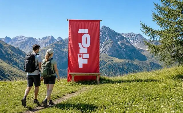 Zwei Wandernde stehen auf einer Almwiese vor einem roten Tirol-Banner mit Blick auf eine alpine Berglandschaft. Bild steht für Natur, Tourismus und Bergerlebnis in Tirol.