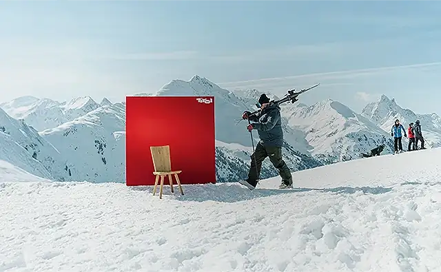 Szene aus der zielgruppenspezifischen Werbekampagne 'Immer für dich da'. Skifahrer, mit Skiern auf seiner Schulter der auf der Skipiste an einer Werbewand von 'Tirol' vorbeigeht, vor der ein traditioneller Holzsessel steht. Im Hintergrund sieht man weitere Skifahrer rasten und ein wunderschönes Bergpanorama.
