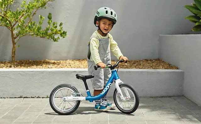 Kind mit Helm steht mit einem blauen woom-Laufrad in einem sonnigen Innenhof. Bild steht für Kinderfahrräder, sichere Mobilität und spielerisches Lernen beim Radfahren.