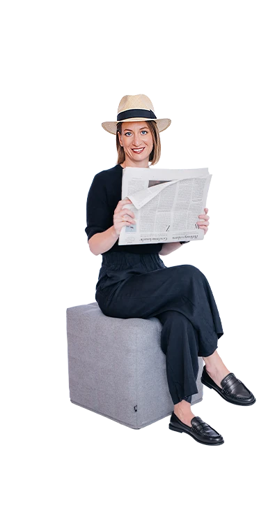 Barbara Khayat-Platzl sitzt mit übergeschlagenen Beinen und Zeitung in der Hand lächelnd auf einem Hocker - entspannt, stilvoll und immer gut informiert.