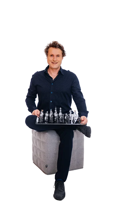 Dr. Gereon Friederes sitzt entspannt auf einem grauen Hocker und hält ein Schachbrett mit transparenten Figuren - strategisch, fokussiert und mit einem Lächeln.
