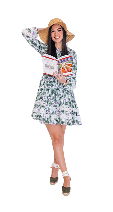 Giselle Kaufmann trägt ein geblümtes Sommerkleid und einen Sonnenhut, während sie mit einem Madrid-Reiseführer lächelnd Fernweh weckt.
