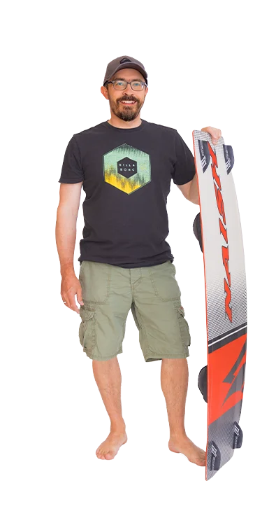 Gerhard Schüller steht barfuss, trägt ein T-Shirt mit Surfmotiv und hält ein Kiteboard - sportlich, entspannt und bereit für Wind und Wellen.