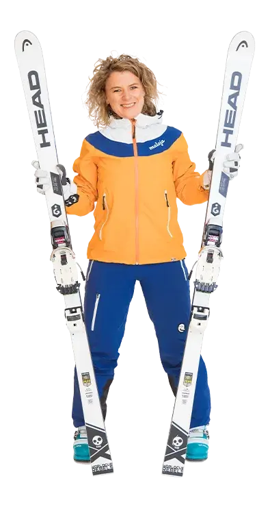 Heidi Thyri steht in kompletter Skimontur mit orange-blauer Jacke und hält zwei weisse Skier fest in den Händen - bereit für die Piste und jede Menge Schneeabenteuer.