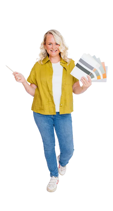 Kerstin Hofer steht mit Farbfächer und Pinsel in der Hand und lächelt begeistert in die Kamera - kreativ, lebensfroh und voller Gestaltungslust.