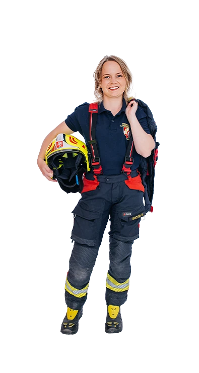 Nadine Buchta trägt eine dunkelblaue Feuerwehr-Einsatzhose mit roten Hosenträgern und gelben Reflexstreifen, den Feuerwehrhelm unter dem Arm und die Einsatzjacke über der Schulter - mit Leidenschaft bei der freiwilligen Feuerwehr.