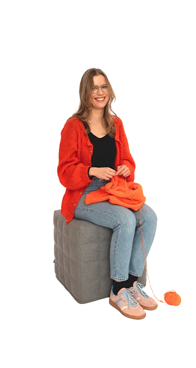 Polina Yaroshchuk sitzt mit oranger Strickjacke und Wolle auf einem Hocker, das Strickzeug in der Hand - kreativ, konzentriert und handwerklich geschickt.